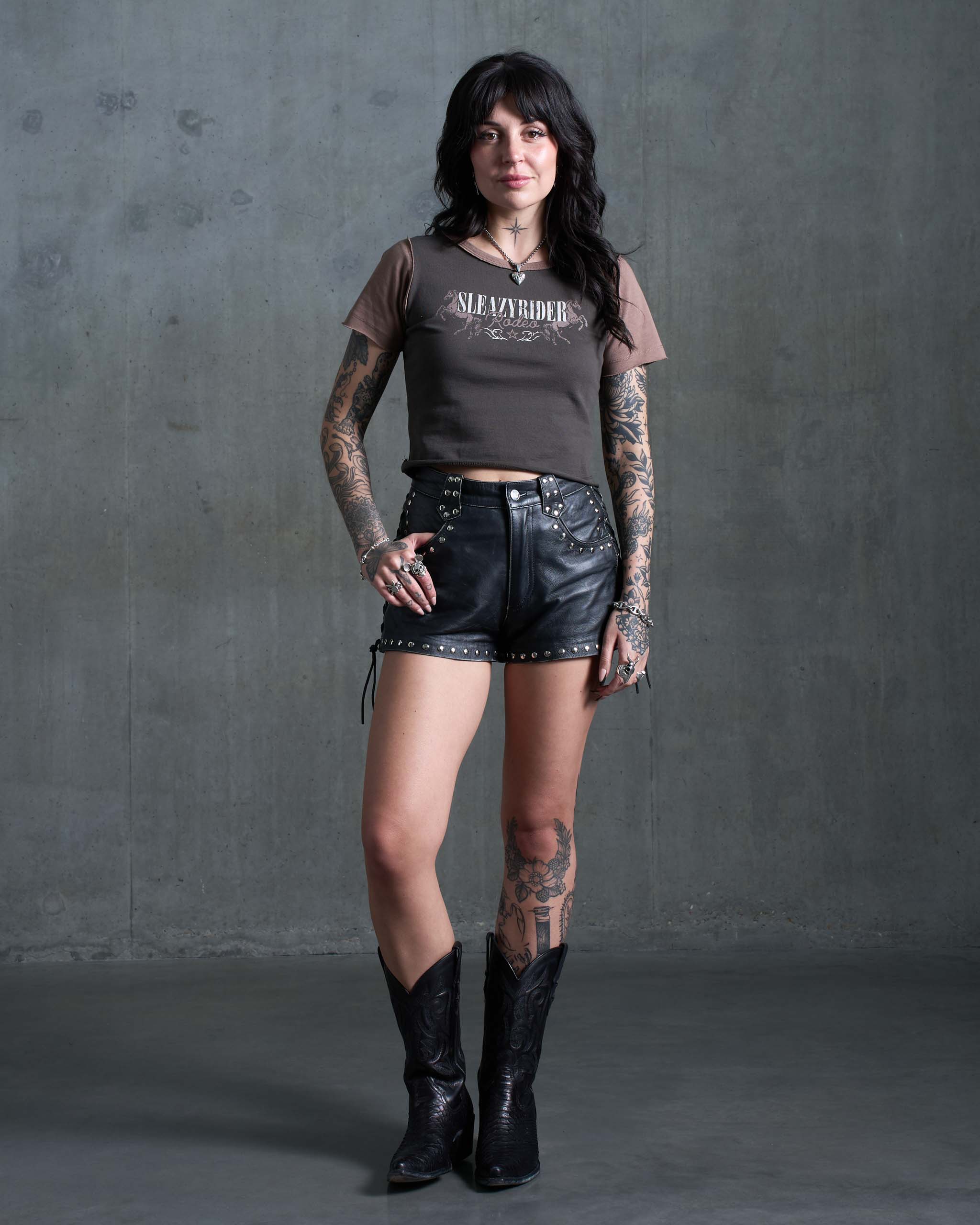 An alternative woman wearing a Sleazy Rider dark western baby t-shirt, with studded shorts and cowboy boots.