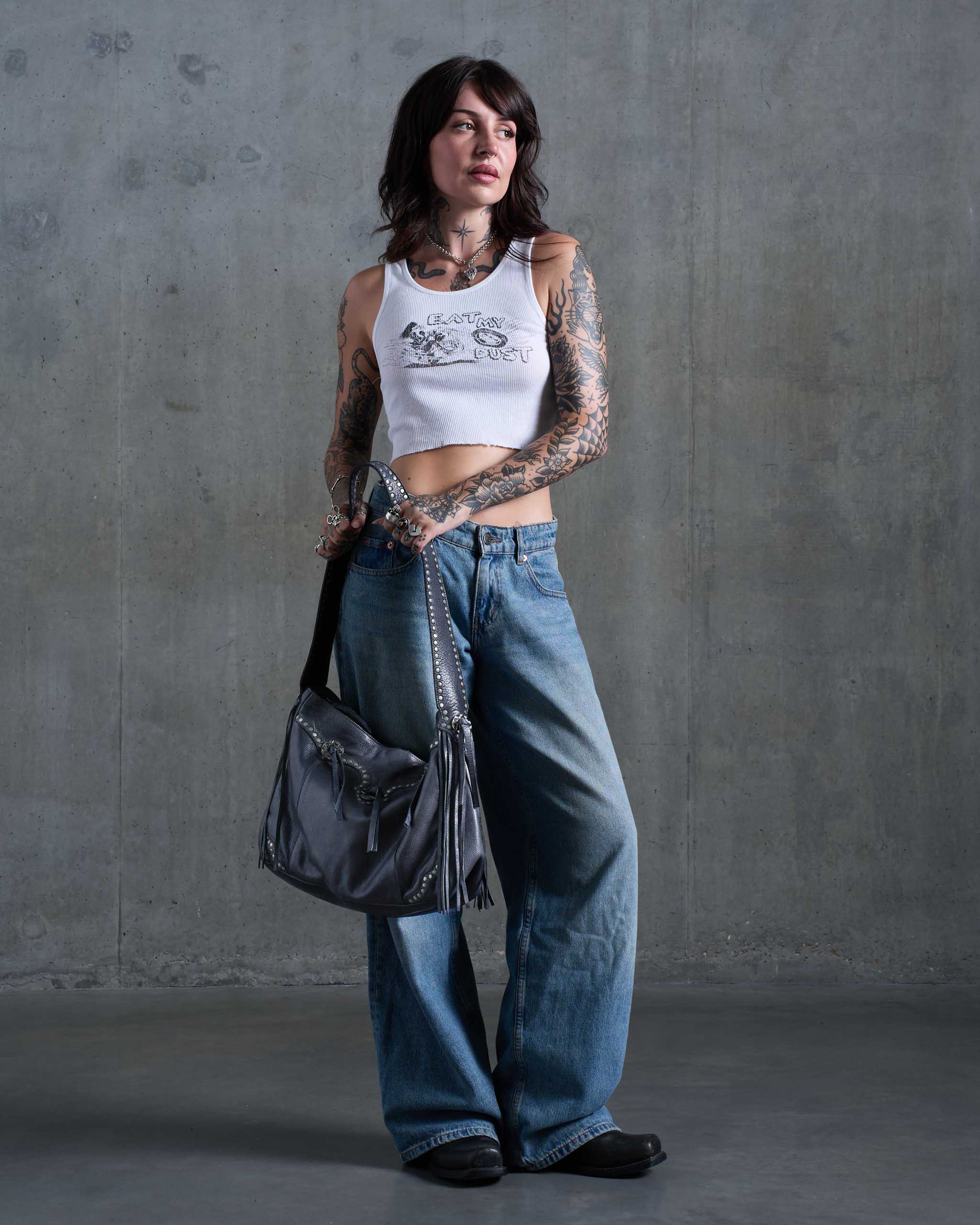 A tattooed model stands against a textured gray concrete wall holding a grey western bag with studs, fringe and conchos.