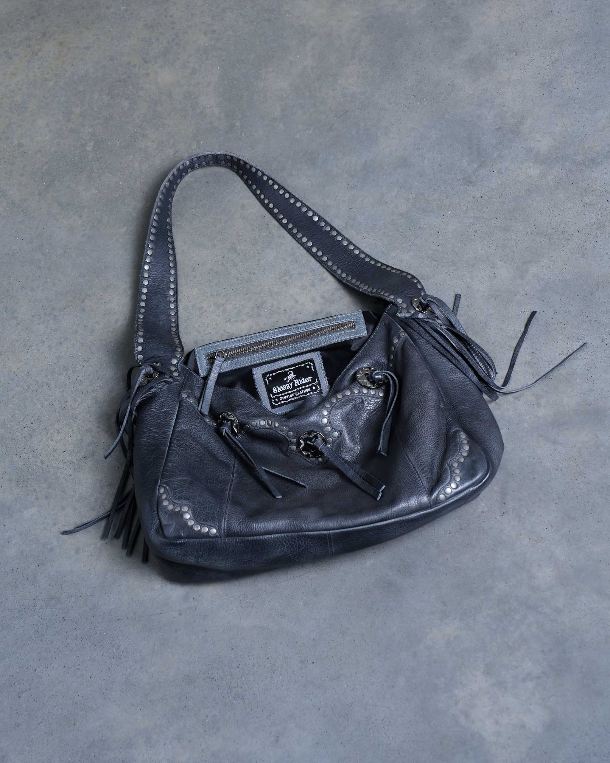 A dark gray western leather shoulder bag with fringe details, silver studs and conchos, displayed on a textured gray concrete surface.