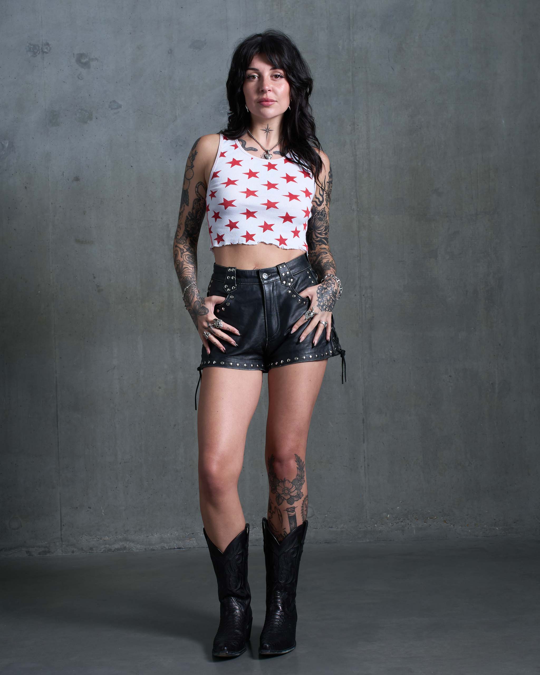 A tattooed woman wearing a tank top with stars all over print, studded leather shorts, and cowboy boots.