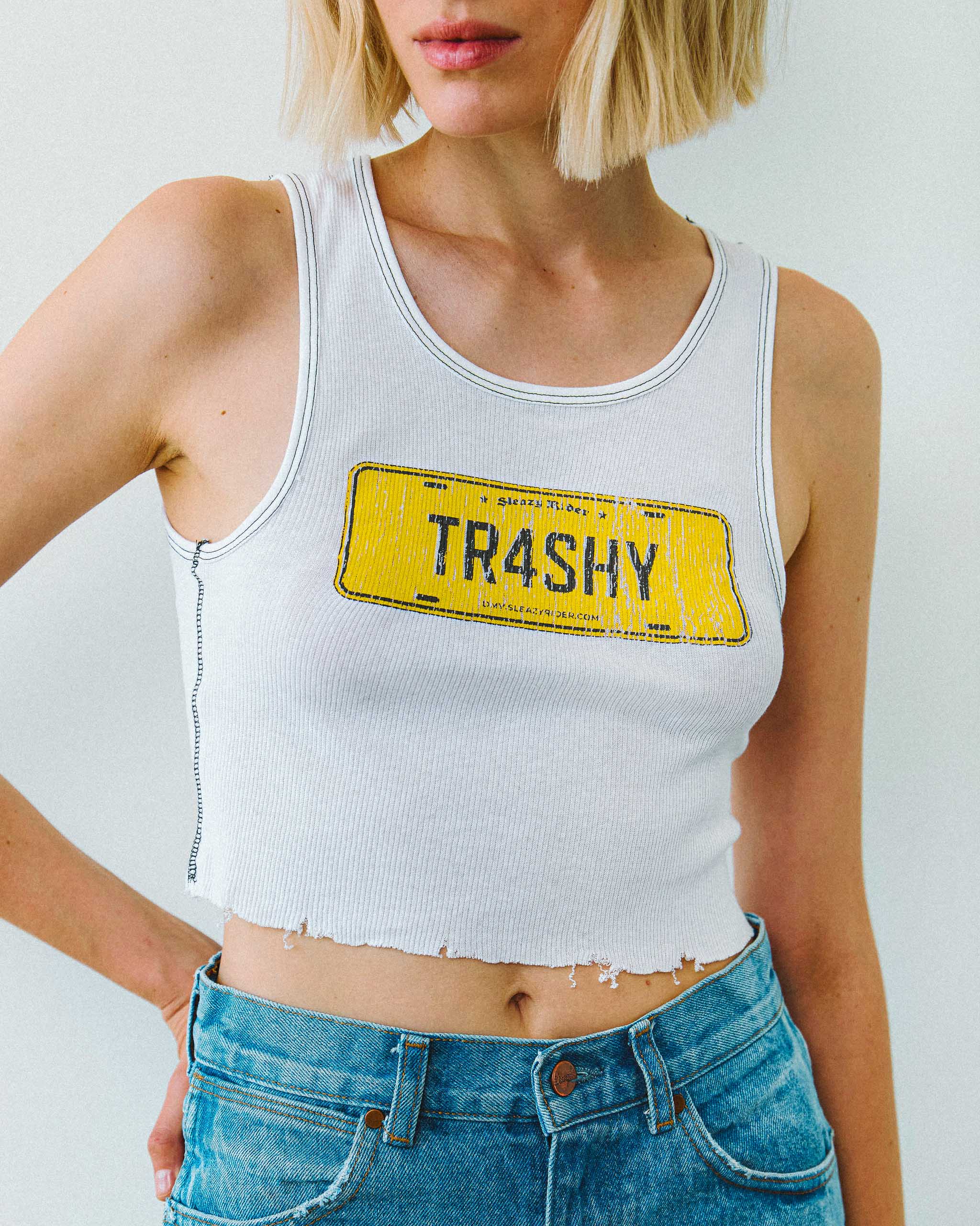A female model wearing a white "TRASHY" graphic tank top with contrast stitching, standing against a plain background.