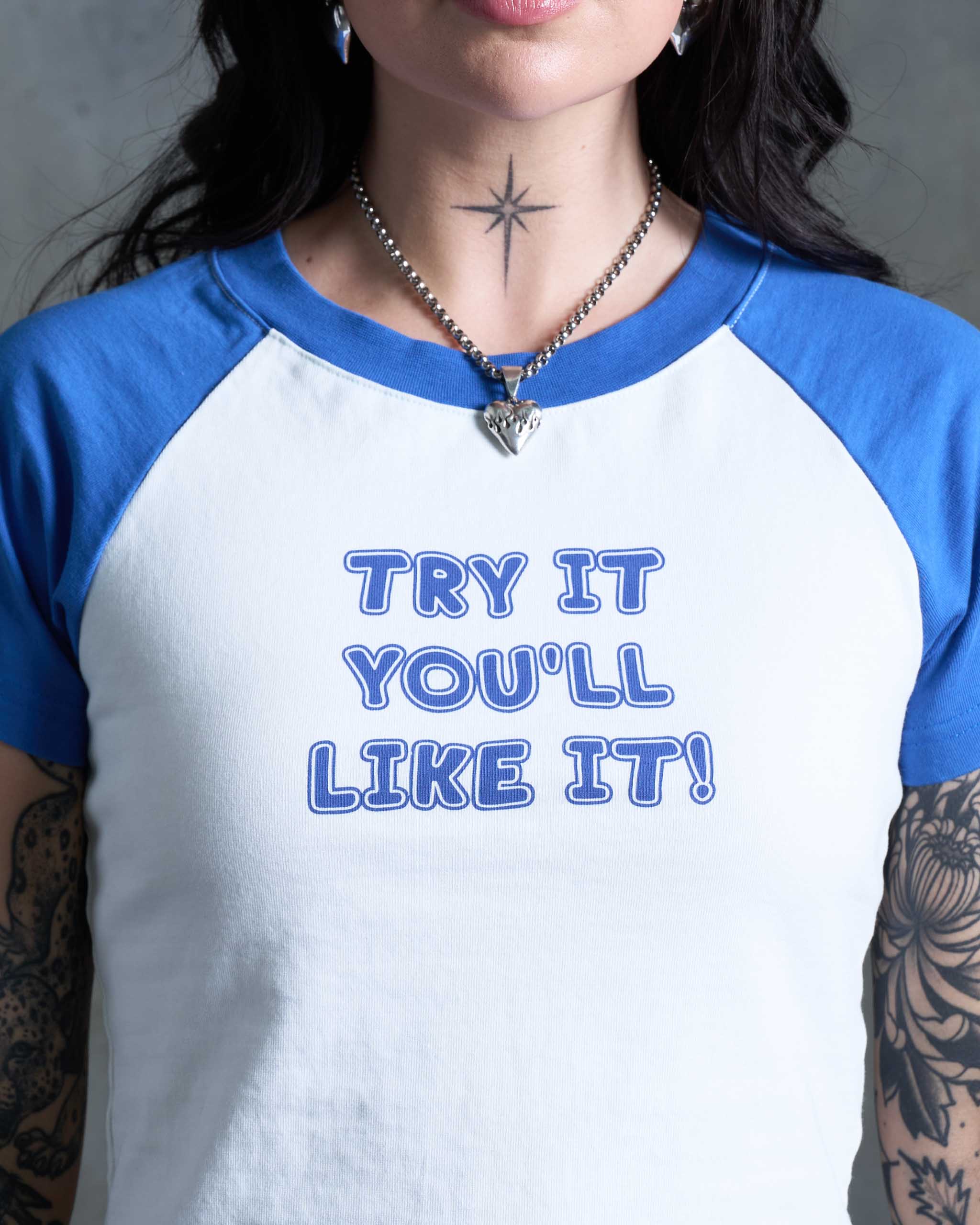 Tattooed woman wearing a blue and white vintage style "Try it you'll like it!" slogan t-shirt, standing against a concrete wall.
