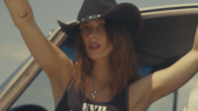 A dark western cinematic video featuring models wearing Sleazy Rider tank tops and cowboy hats, posing in front of vintage Corvettes and Harley Davidsons.